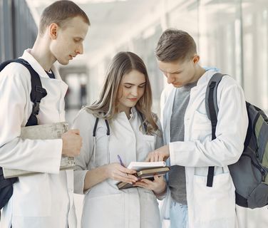 Three medical students in white coats with backpacks discuss notes in a bright hallway.
