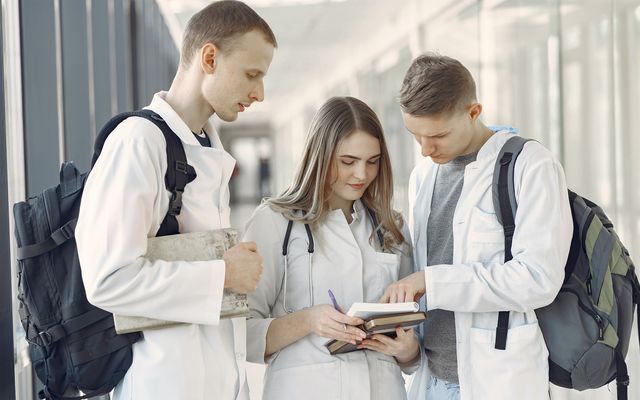 Three medical students in white coats with backpacks discuss notes in a bright hallway.