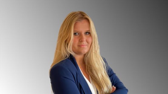 Portrait einer Frau mit langen blonden Haaren im blauen Blazer vor grauem Verlaufshintergrund.