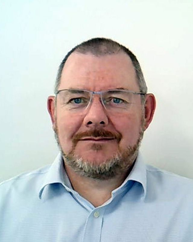 Portrait of a man with glasses and a beard in a light blue shirt, plain background.