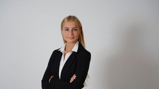 Woman in business attire standing with arms crossed against a plain background, speaker portrait.