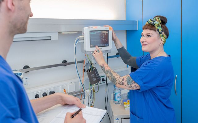 Nurse in blue scrubs operates hospital monitor; another person writes on a chart nearby.