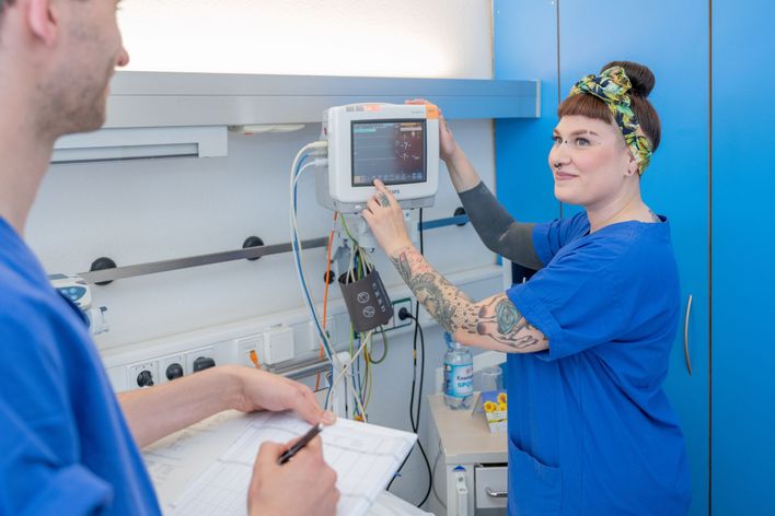 Nurse in blue scrubs operates hospital monitor; another person writes on a chart nearby.