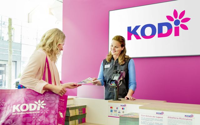 Two women at KODi store counter, one with KODi bag, pink branding, KODi DER HAUSHALTDISCOUNTER.