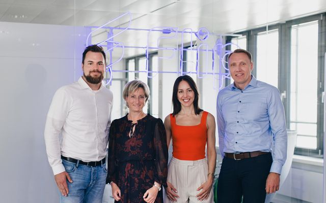 Four people standing in front of a neon Flip sign, office environment, no UI or app visible.
