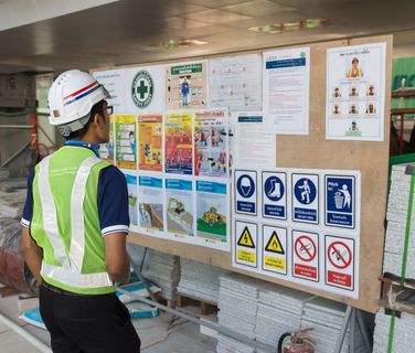 Construction worker in safety gear reads a site safety board with Thai instructions and warning signs.