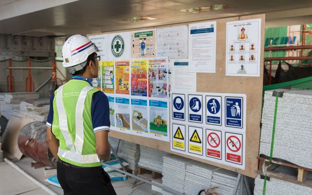 Construction worker in safety gear reads a site safety board with Thai instructions and warning signs.
