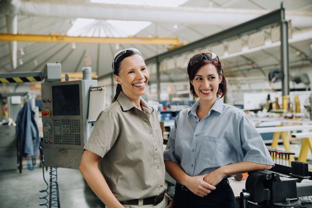 Two smiling manufacturing workers in uniforms and safety glasses in a bright factory setting.