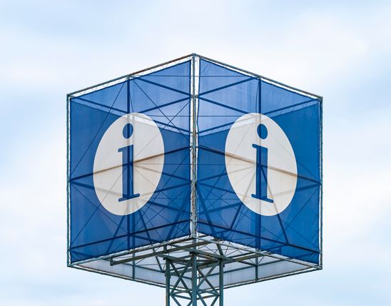 Large blue cube sign with white i for information, outdoors against a cloudy sky.
