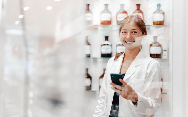 Female pharmacist in white coat smiling, holding phone, standing by medicine bottles in pharmacy.