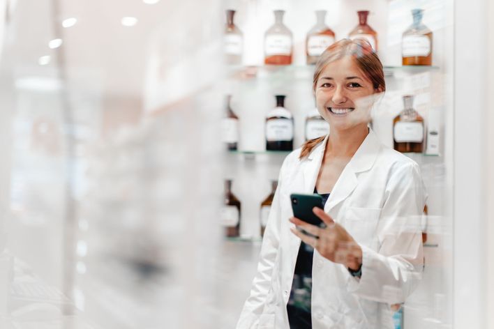 Female pharmacist in white coat smiling, holding phone, standing by medicine bottles in pharmacy.
