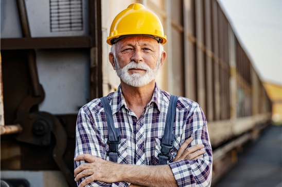 Older frontline worker in a hard hat and plaid shirt standing by industrial equipment outdoors.
