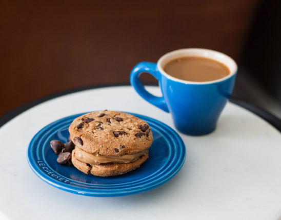 Chocolate chip cookie sandwich with chocolate chips on a blue plate, next to a blue cup of coffee.