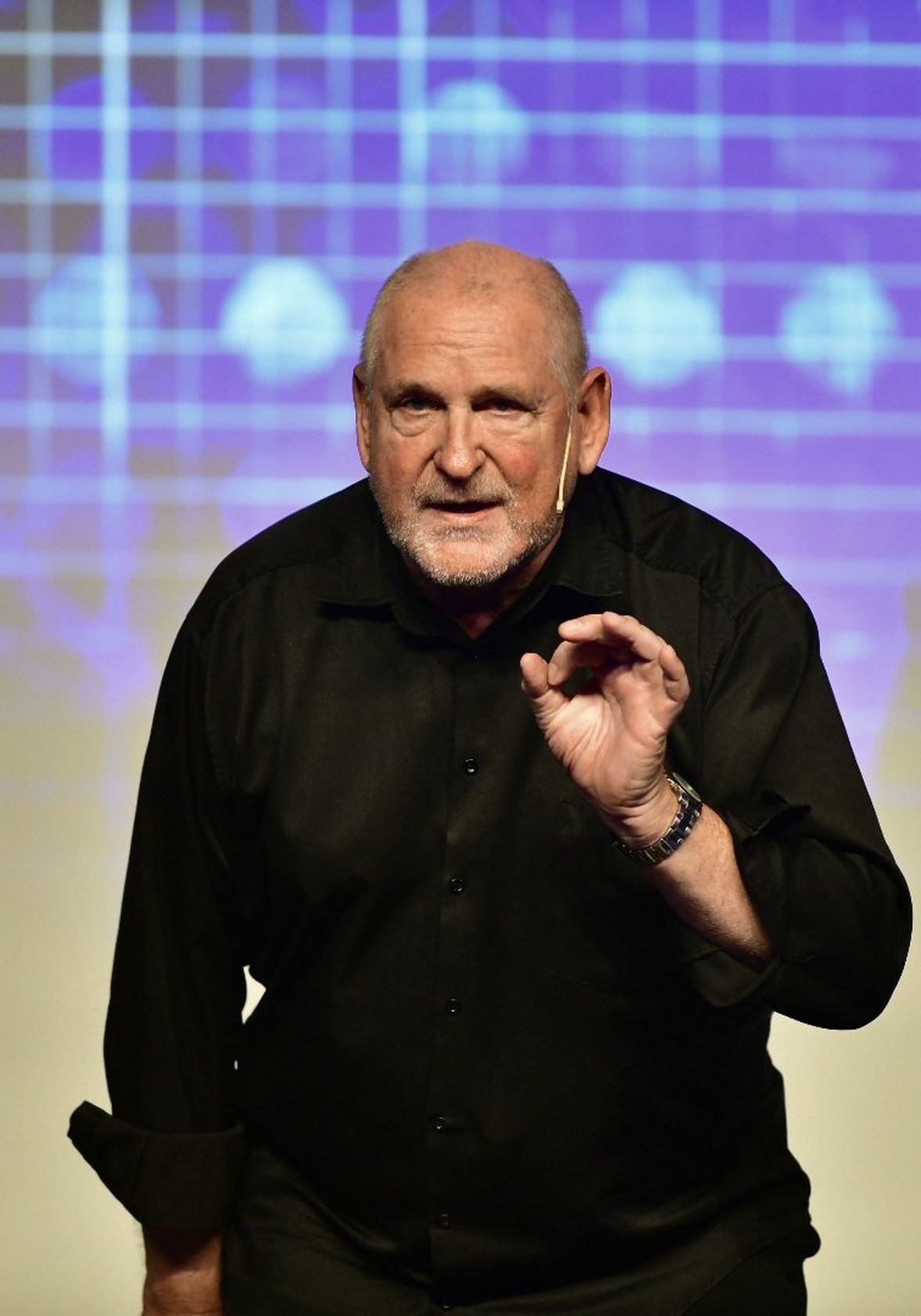 A man in black shirt speaks on stage with a digital grid background, no visible text.