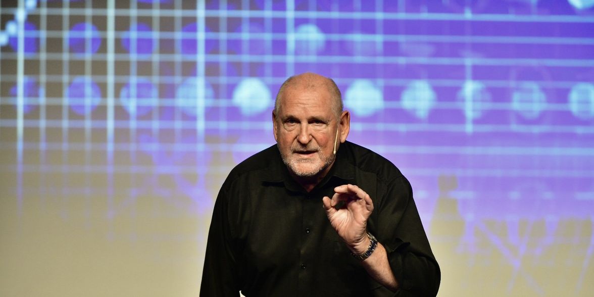 A man in black shirt speaks on stage with a digital grid background, no visible text.