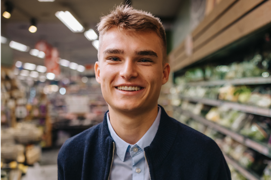 Young man smiling in a grocery store aisle, shelves stocked with produce in the background.