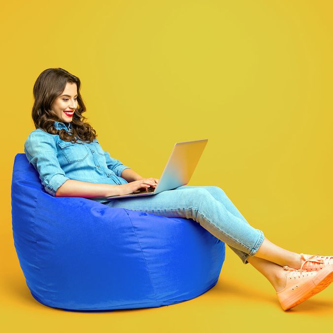 Woman in casual clothes using laptop on blue beanbag, yellow background, modern remote work.