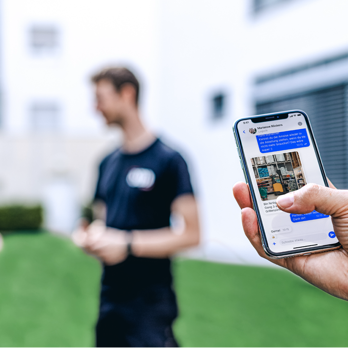 Hand holding phone with German text, blurred people in background.