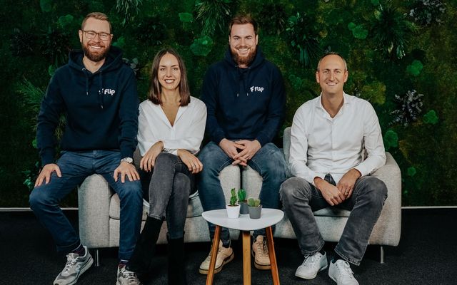 Four people sit on a couch in front of a green wall, two wear Flip hoodies, smiling at camera.