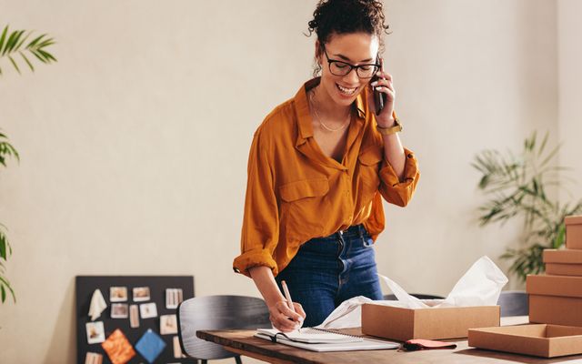 Woman packing boxes and talking on phone in home office, representing new HR roles and flexibility.