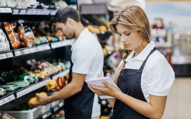 Woman in supermarket aisle taking notes, man stocking produce, both in uniforms, grocery setting.