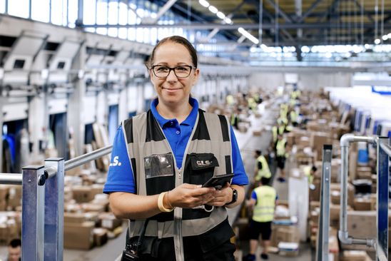 Warehouse worker in GLS uniform holding phone, standing in busy logistics center, smiling at camera.