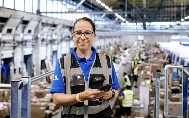 Worker in a busy warehouse holding a phone, surrounded by packages and colleagues.