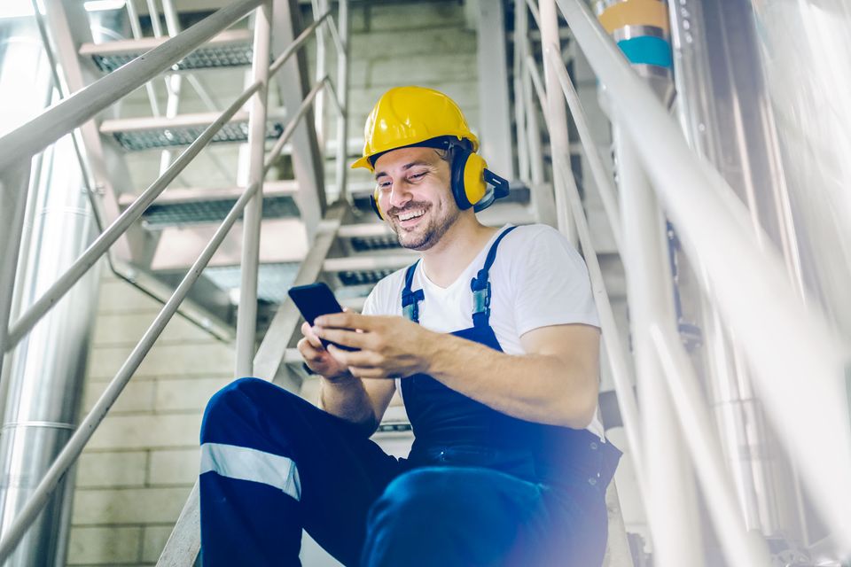 Smiling worker in helmet uses smartphone, symbolizing Employee App, Mitarbeiterplattform, HR-Software.