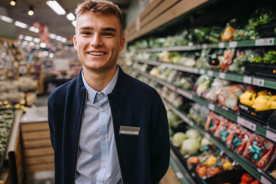Smiling supermarket worker in produce aisle, wearing a name tag, fresh vegetables in background.