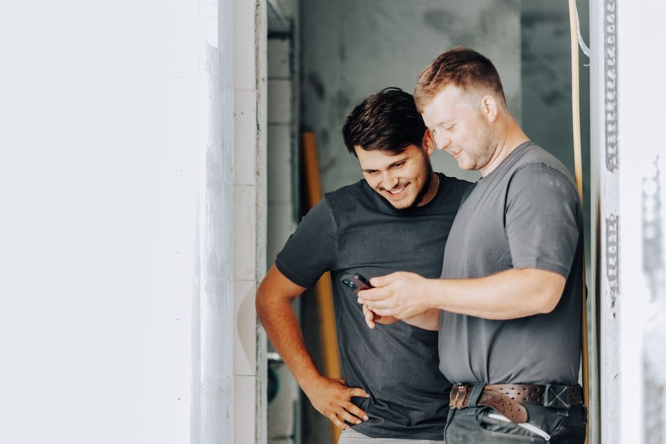 Two men smiling and looking at a phone in a bright room.