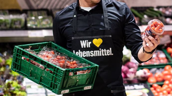 Supermarktmitarbeiter mit Schürze Wir ♥ Lebensmittel hält Kiste und Becher mit Tomaten.