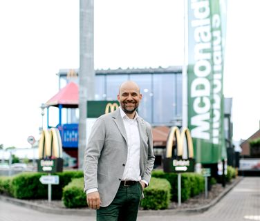A man in a suit smiles in front of a McDonalds restaurant and green McDonalds flag, Einfahrt.
