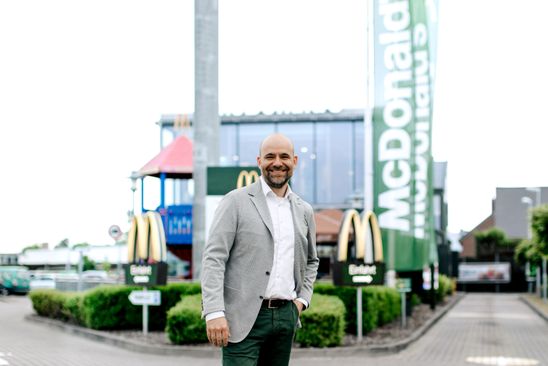 A man in a suit smiles in front of a McDonalds restaurant and green McDonalds flag, Einfahrt.