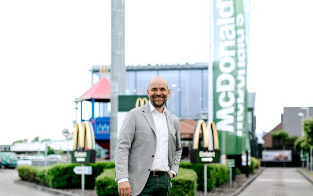 A man in a suit smiles in front of a McDonalds restaurant and green McDonalds flag, Einfahrt.