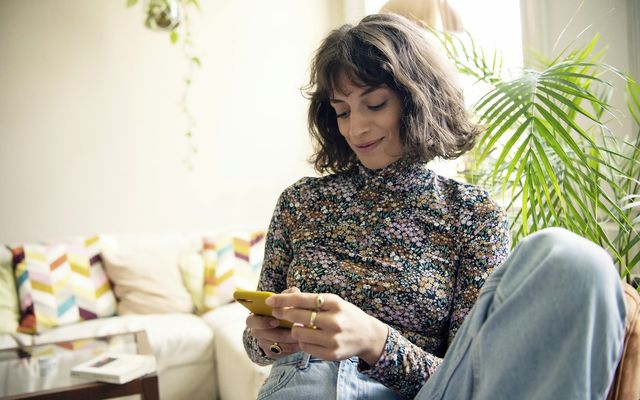 Woman sitting on sofa at home, smiling at yellow smartphone, with colorful pillows and plants.