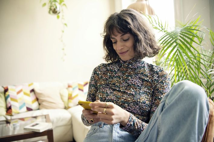 Woman sitting on sofa at home, smiling at yellow smartphone, with colorful pillows and plants.