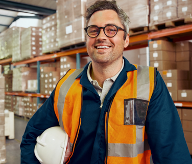 Warehouse worker in safety vest smiling, surrounded by boxes; no visible software interface or text.