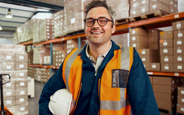 Warehouse worker in safety vest smiling, surrounded by boxes; no visible software interface or text.
