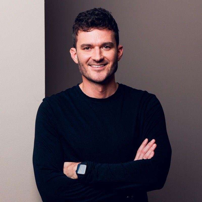 Portrait of a smiling man in a black shirt with arms crossed, standing against a neutral background.
