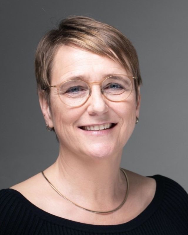 Portrait of a smiling woman with short hair and glasses, wearing a black top and necklace.