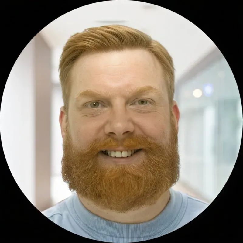 Portrait of a smiling man with red hair and beard, blurred indoor background, circular frame.