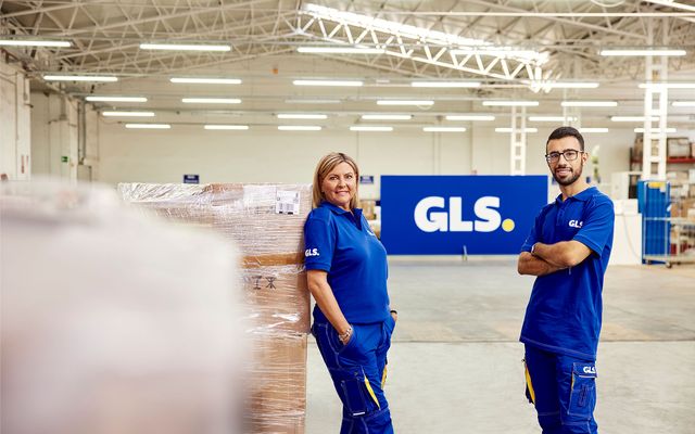 Two GLS employees in blue uniforms stand in a warehouse, large GLS sign in background.