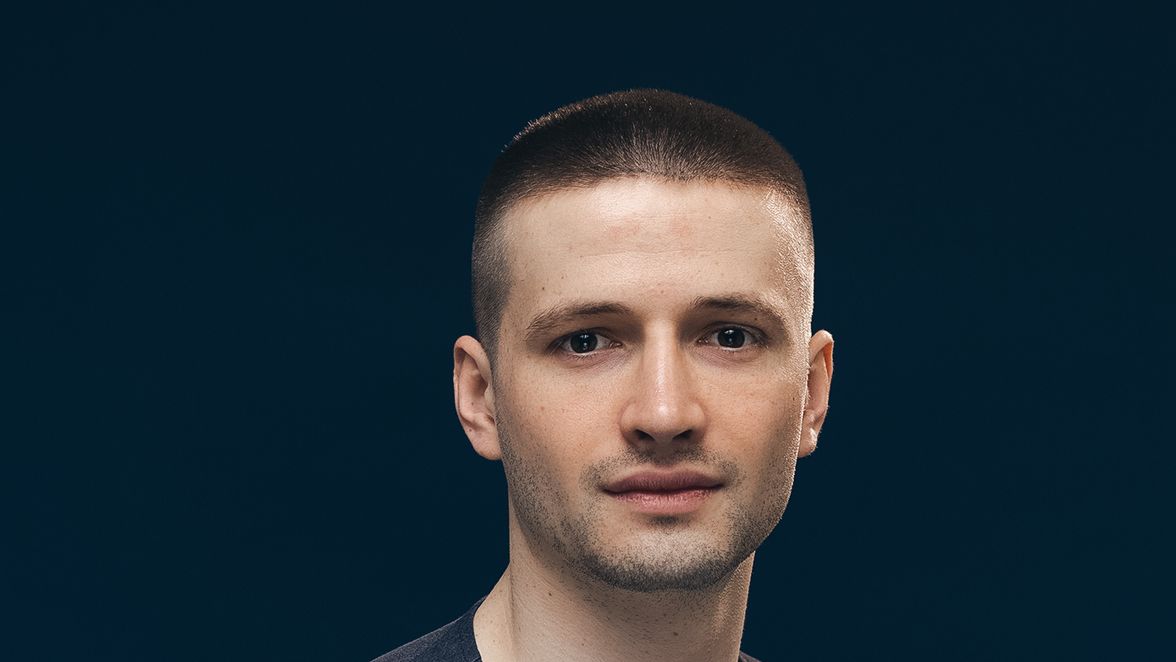 Portrait of a man in a dark t-shirt against a plain dark background, neutral expression.