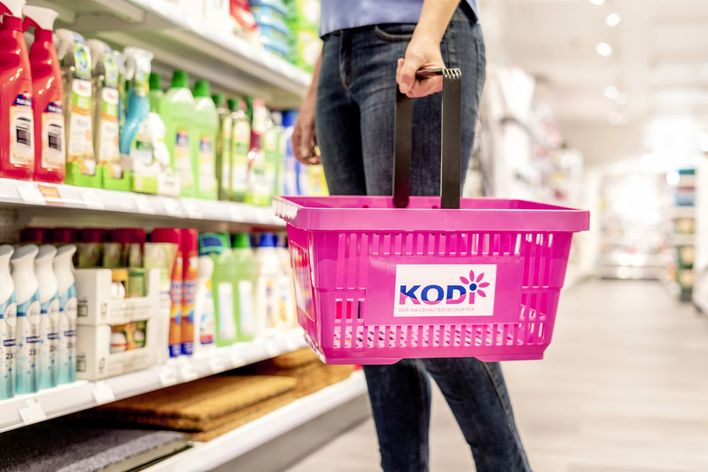 Person holding pink KODi basket in household goods store aisle, shelves with cleaning products.