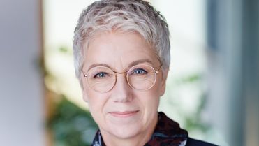 Portrait of an older woman with short gray hair and glasses, wearing a dark top and scarf.