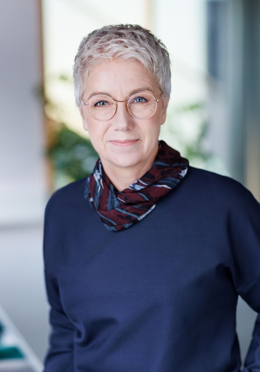 Portrait of an older woman with short gray hair and glasses, wearing a dark top and scarf.