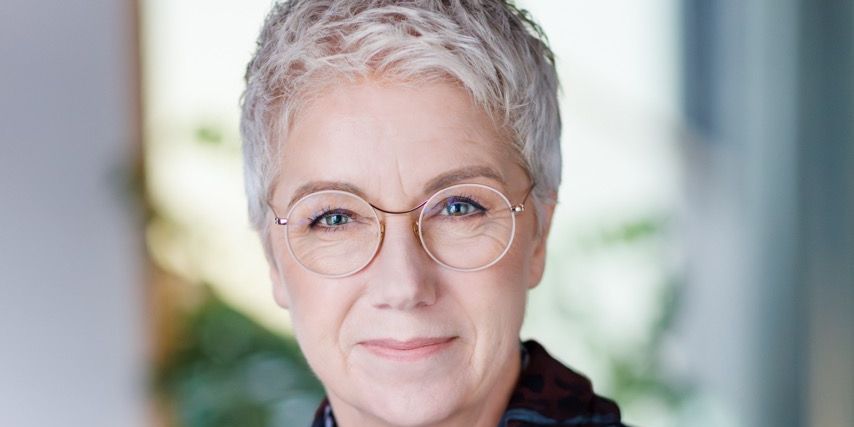 Portrait of an older woman with short gray hair and glasses, wearing a dark top and scarf.