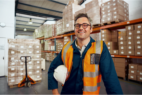 Warehouse worker in safety vest smiling, surrounded by boxes; no software UI or keywords visible.
