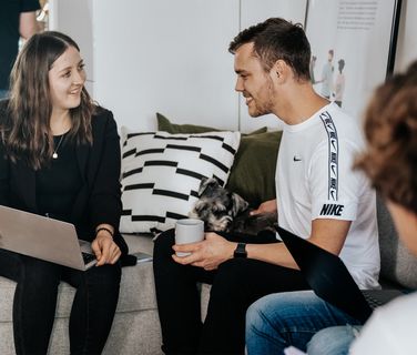 People chatting on a couch with laptops and a dog.