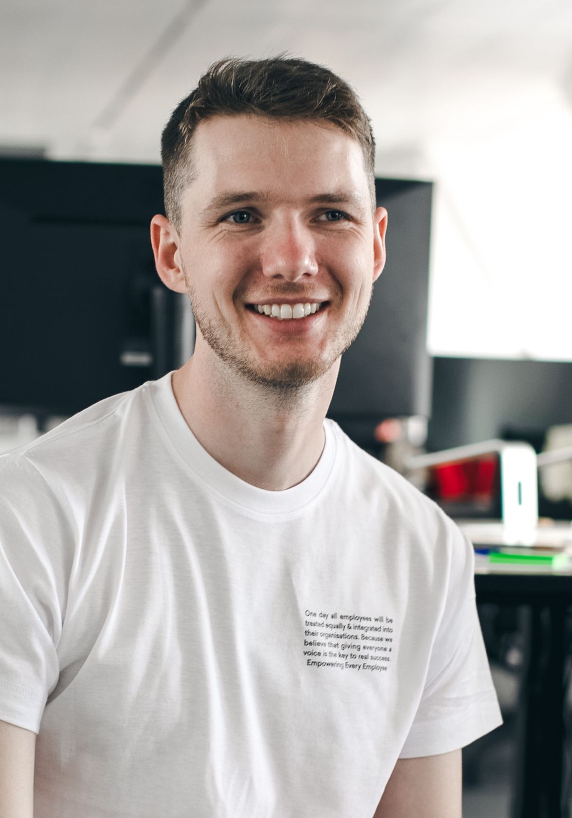 Smiling man in a modern office, wearing a white shirt with English text, testimonial setting.